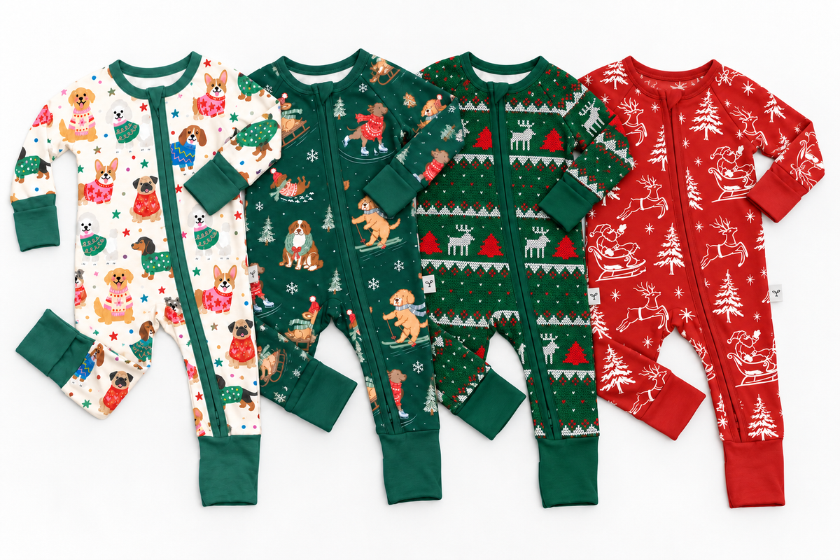 Four baby long-sleeve footless onesies arranged side by side on a white background, each with a different Christmas print: white with dog illustrations, dark green with dog and tree motifs, dark green with reindeer and tree patterns, and red with reindeer and tree patterns.