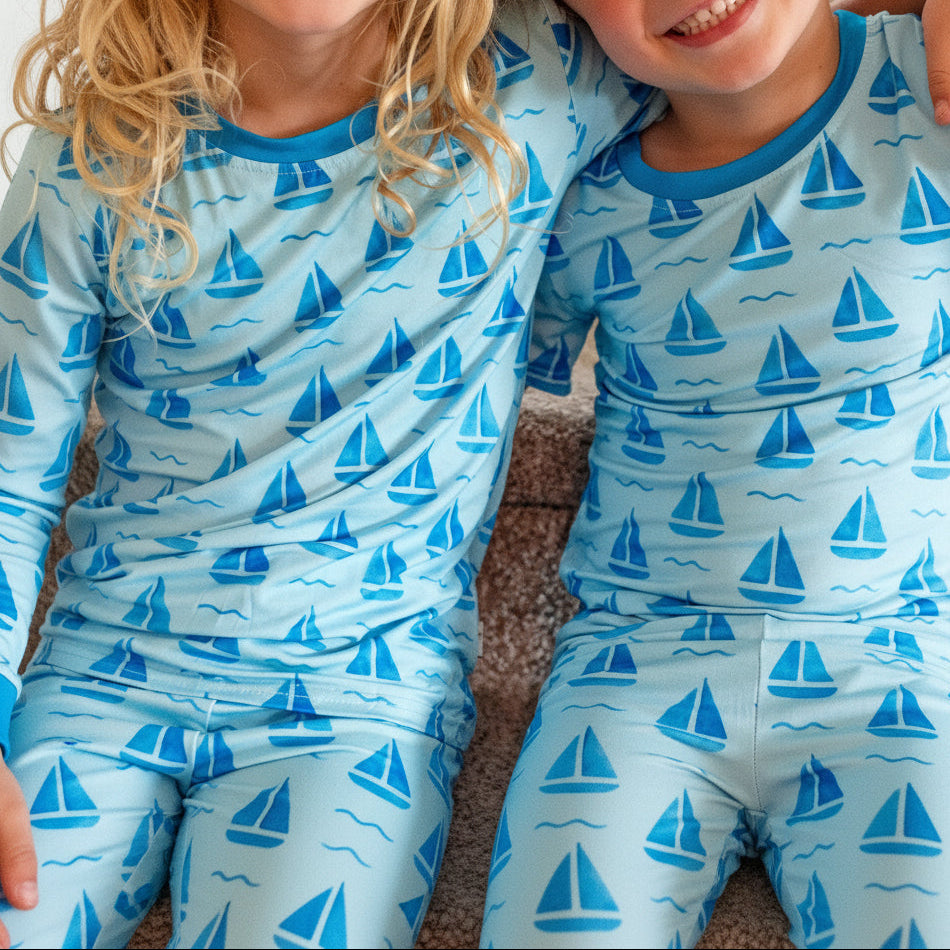 Two children wearing matching light blue long-sleeve pajamas with a repeating blue sailboat pattern, sitting close together on a carpeted floor.