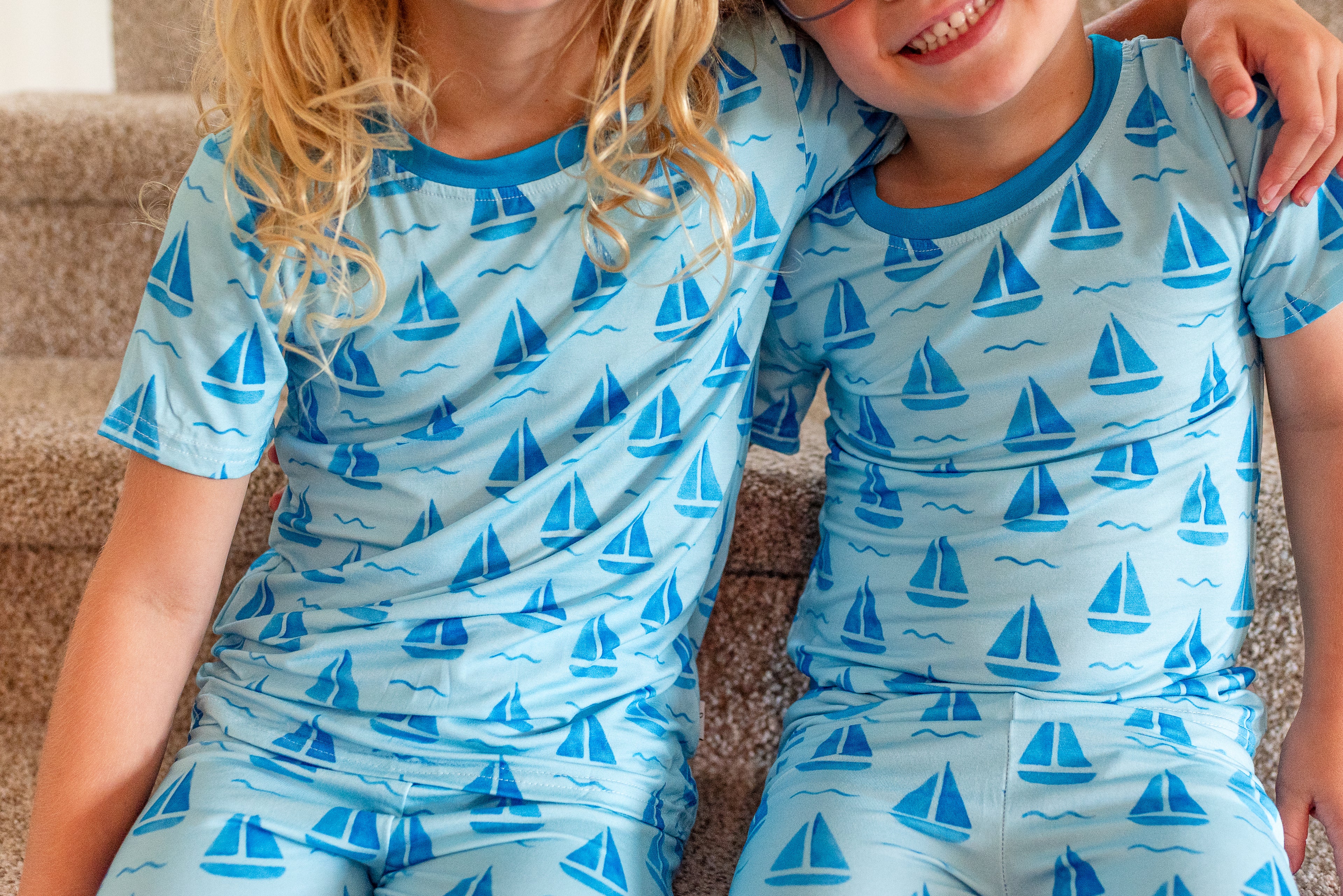 Two children wearing matching light blue pajamas with a blue sailboat pattern, sitting together on a beige carpet.