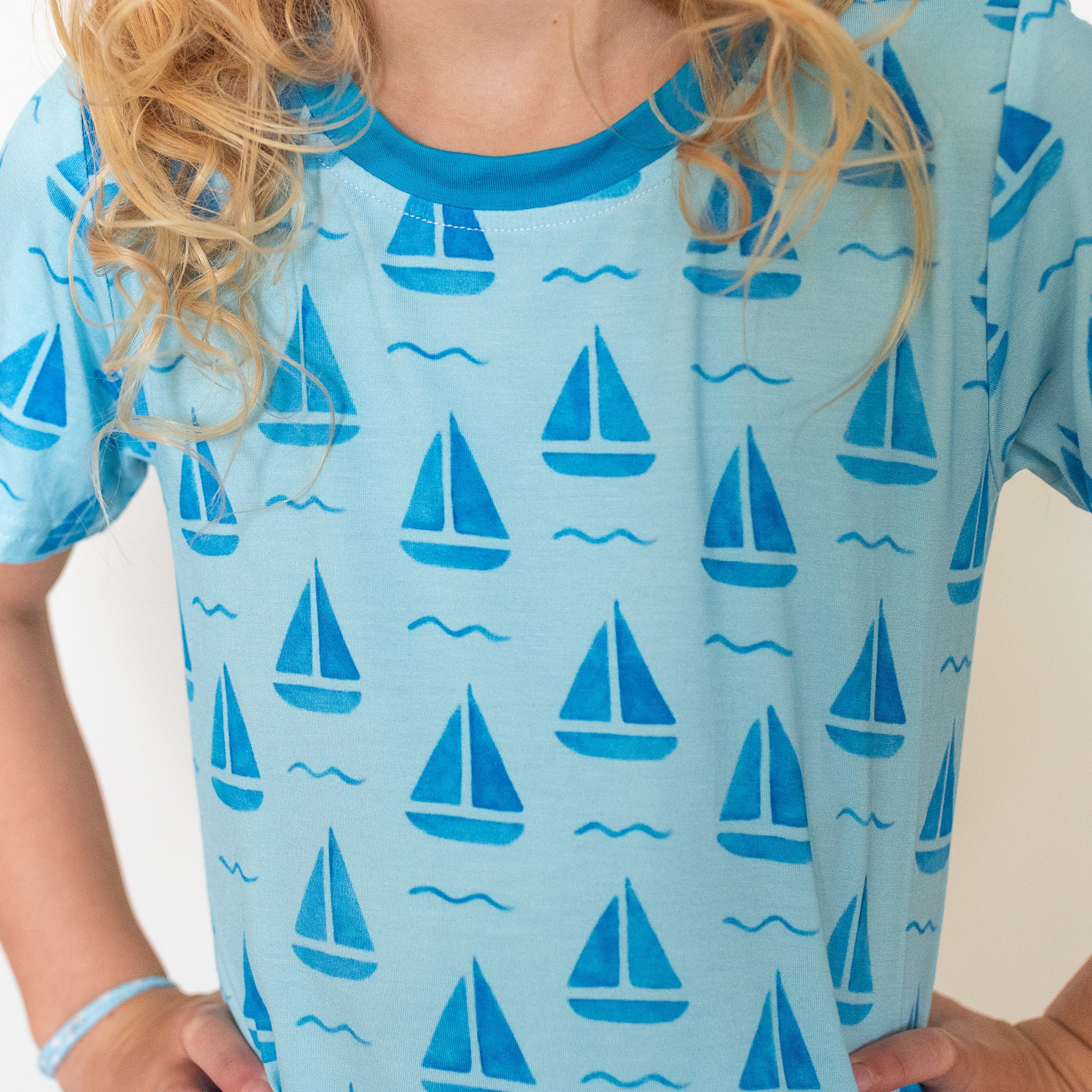 A child wearing a light blue short-sleeve t-shirt with a repeating pattern of blue sailboats and small wavy lines, photographed against a plain light background.